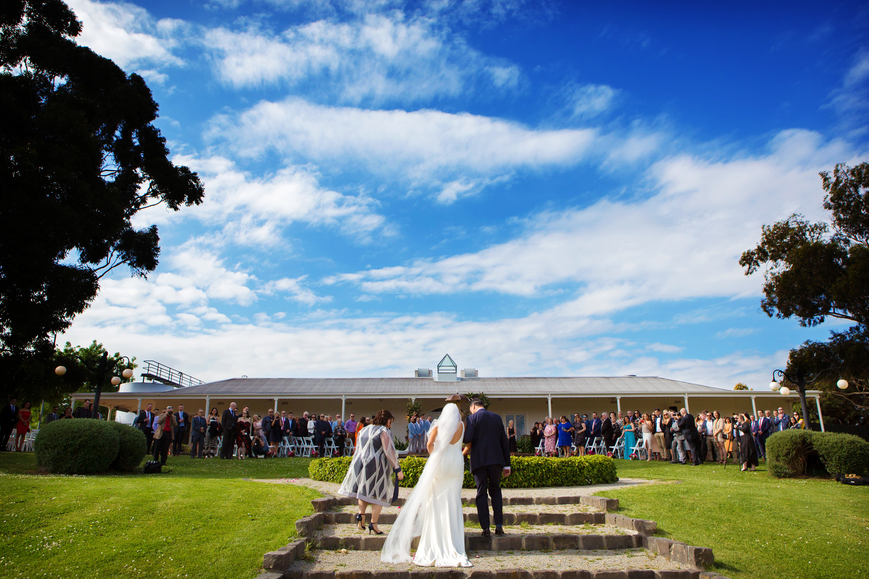 Wide shot of outdoor wedding ceremony. A bride in white gown and groom walk up stone steps at a Yarra Valley style venue under a dynamic blue sky, with many seated guests watching