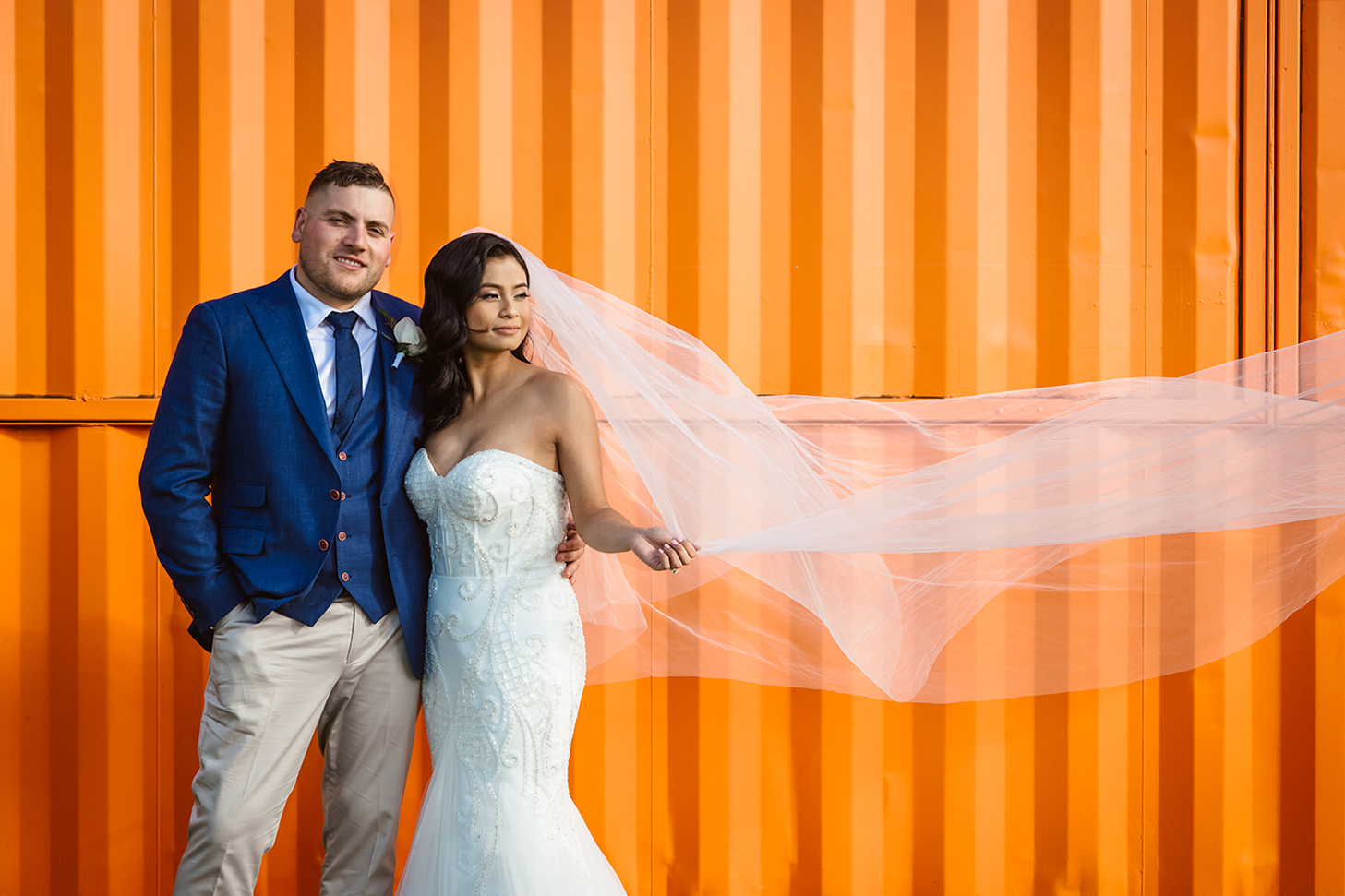 Fun and modern urban wedding portrait. A bride in an intricate gown holds her long veil as it blows in the wind, posing by a bright orange shipping container wall with her groom. Distinctive industrial Melbourne style