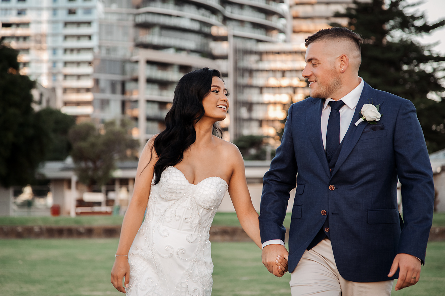 Candid photograph of a bride and groom walking together, holding hands and smiling warmly. Background shows modern inner-city buildings, capturing a natural, joyful moment typical of a contemporary Melbourne wedding