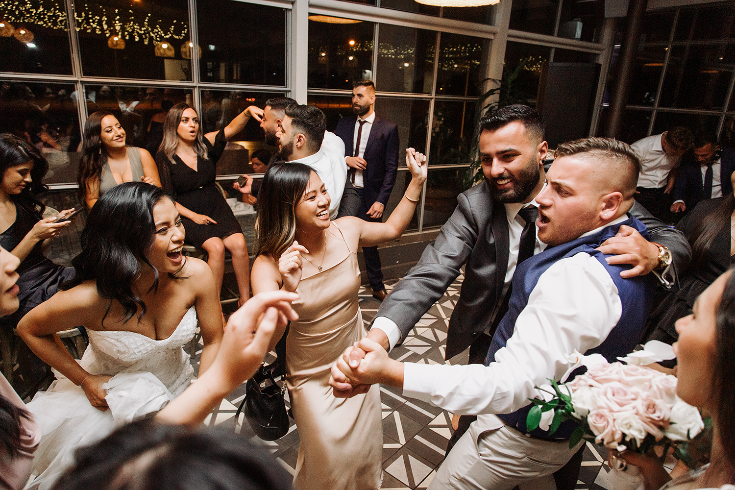 High-energy, candid wedding reception photo. The bride and groom dance joyfully with their friends in a lively indoor party atmosphere. A fun Melbourne wedding celebration with diversely filled dance floor and warm lighting