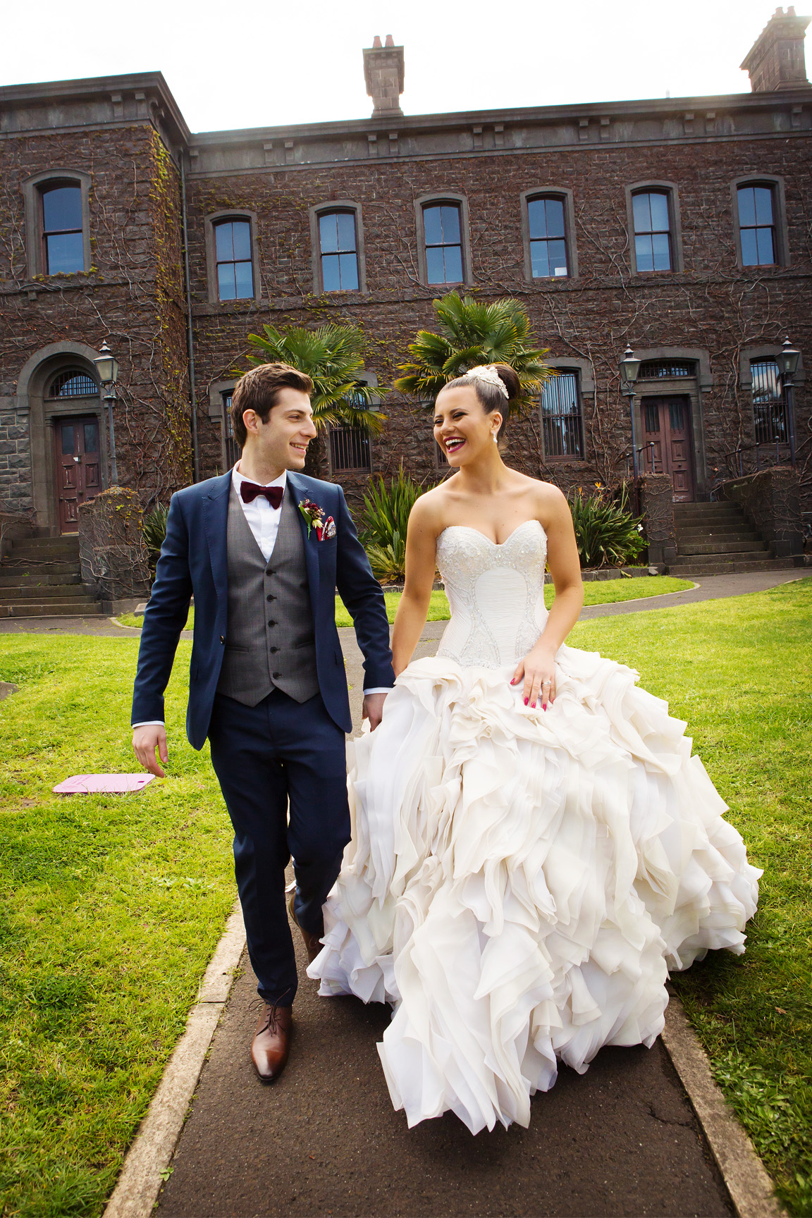 A happy, candid shot of a bride in a voluminous ruffled gown and groom walking and laughing together. They hold hands in front of a grand, historic bluestone building, a classic Melbourne landmark wedding venue covered in ivy