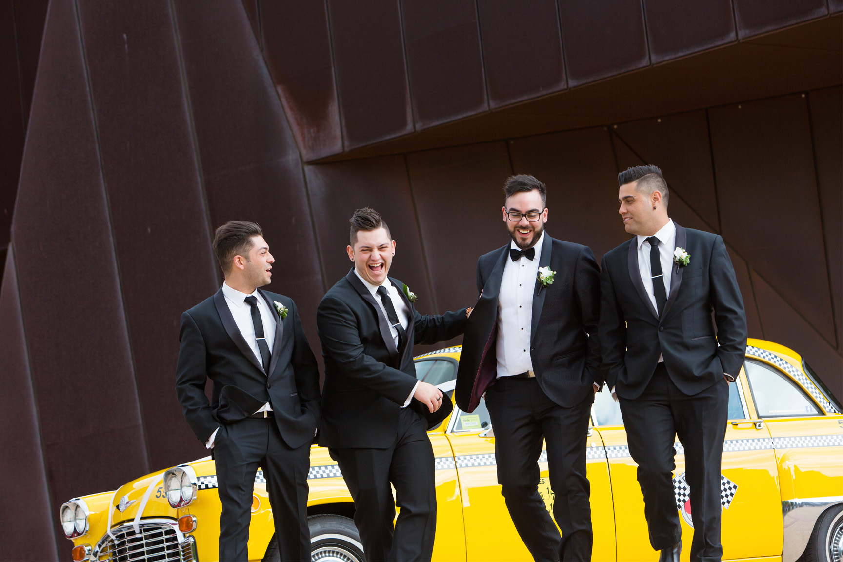 Fun group portrait of four groomsmen in black tuxedos posing and laughing by a vintage yellow checker taxi cab. Background features modern architecture, capturing a playful and stylish moment in modern Melbourne wedding photography