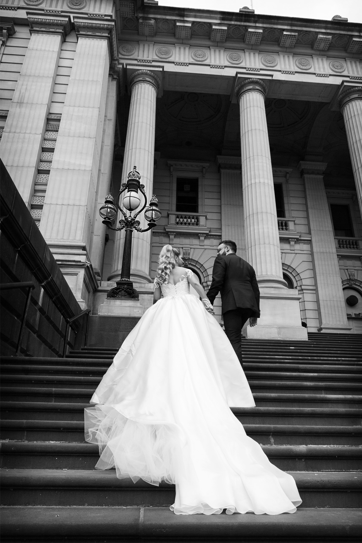 Dramatic, black and white editorial wedding photograph. Shot from a low angle looking up, a bride in a grand gown with a long train and groom ascend majestic stone steps flanked by large pillars at a historic Melbourne landmark venue