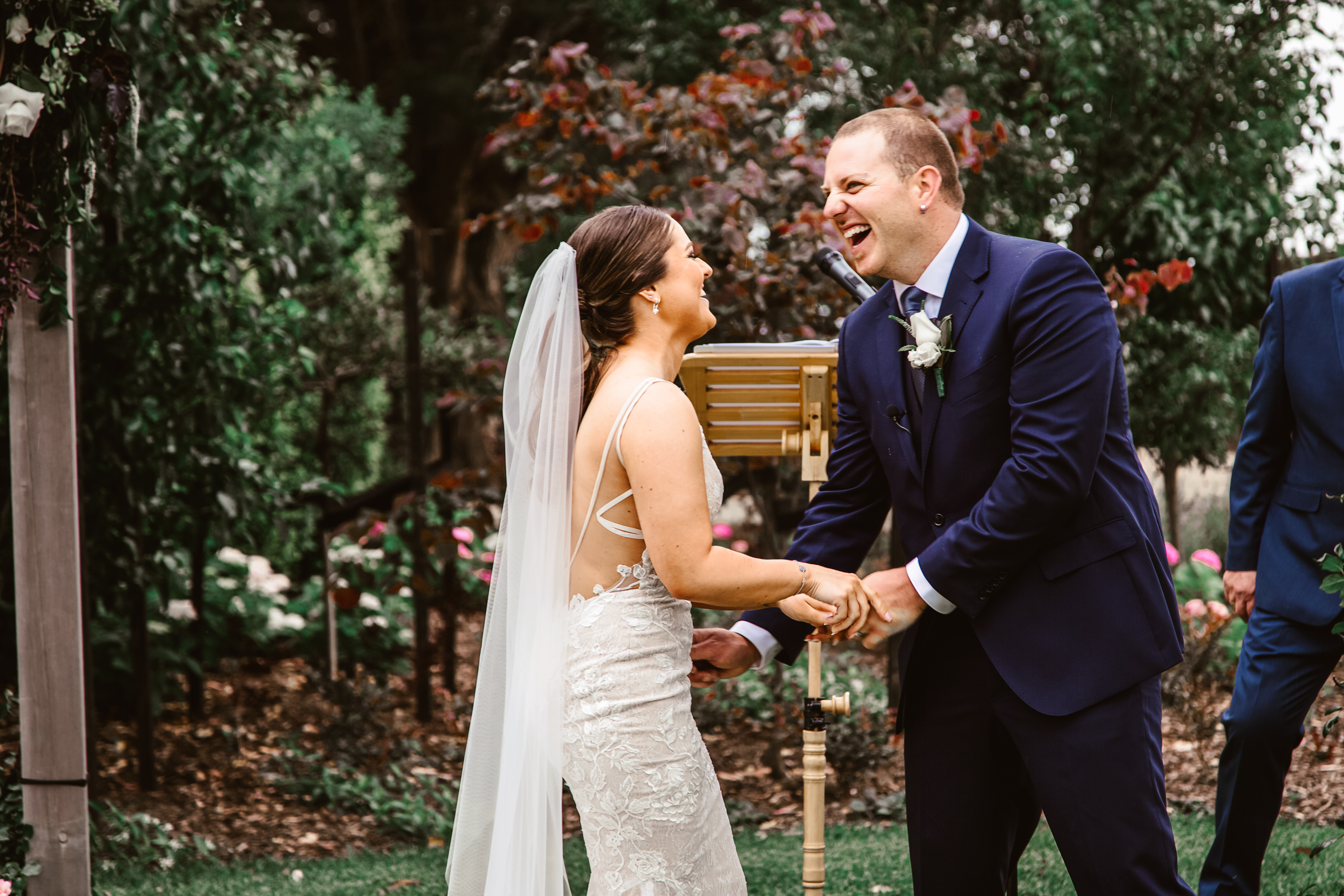 Candid, joyful bride in lace backless dress and groom in navy suit laughing heartily together and holding hands at their outdoor garden wedding altar in Melbourne