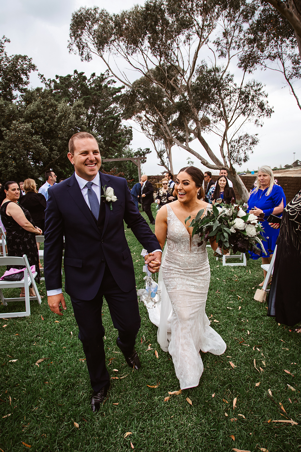 A happy bride and groom walking hand-in-hand down the aisle in a natural garden setting, smiling broadly while guests applaud. Captured by a Mornington Peninsula wedding photographer