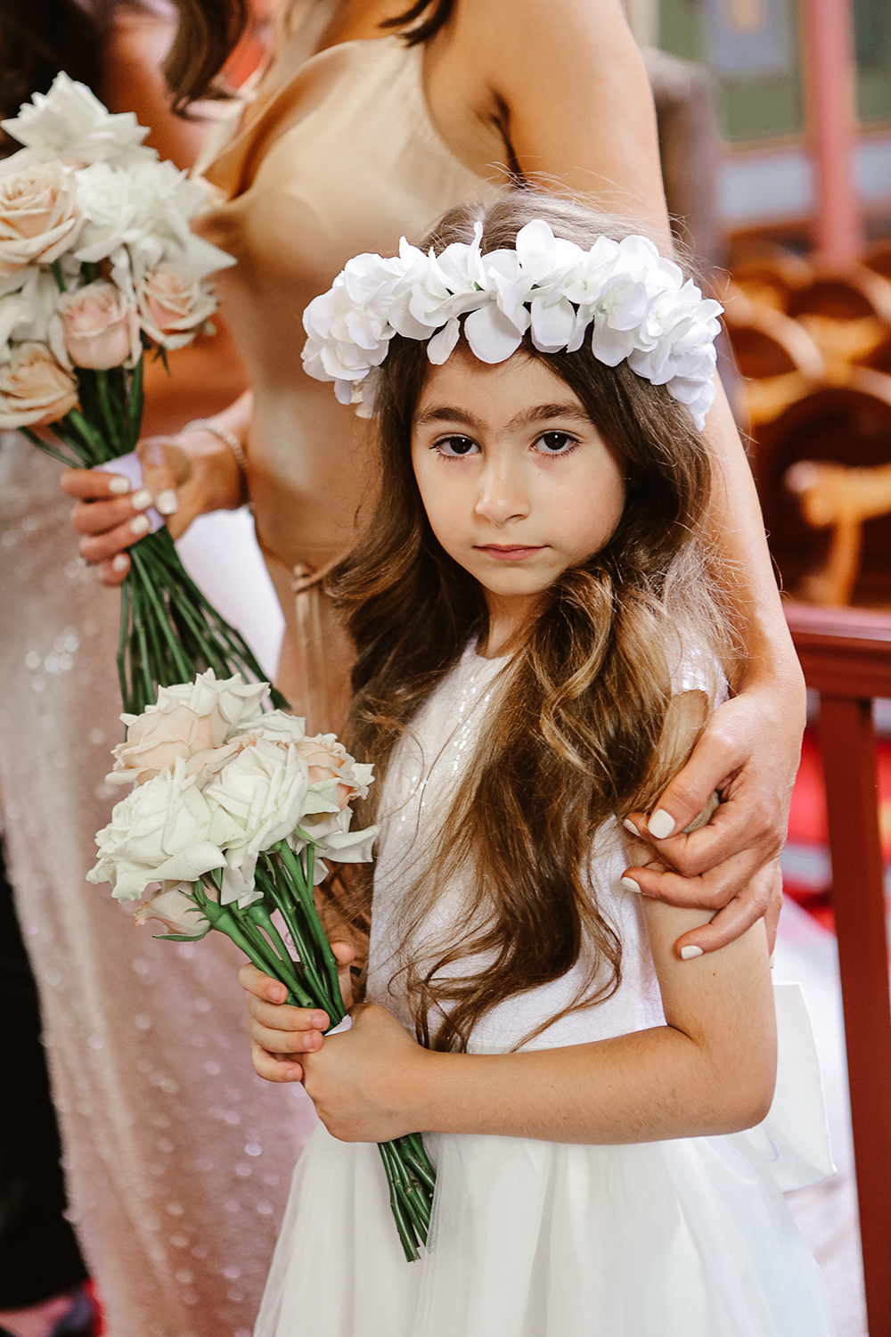 Close-up portrait of a young flower girl with long wavy hair wearing a large white floral crown and holding a white and pink rose bouquet. A sweet, unposed moment during a Melbourne wedding