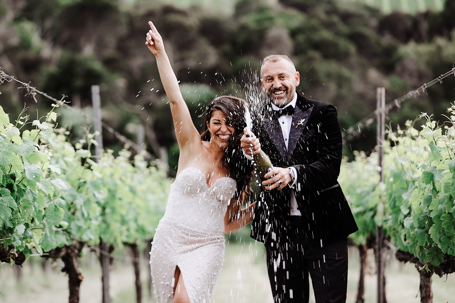 Full-of-life capture of a groom popping a champagne bottle with the cork and liquid spraying, while the bride cheers beside him in a Yarra Valley winery vineyard. Authentic and fun wedding moment