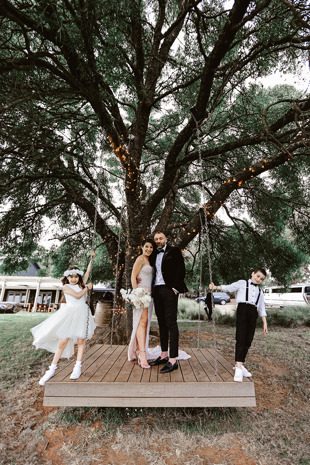 A modern family portrait of a bride and groom posing on a large wooden platform swing with a flower girl and page boy. Captured under a massive, fairy-lit tree at a historic Melbourne property