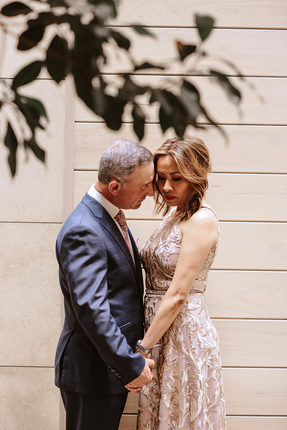 Intimate and natural wedding portrait of a couple with foreheads touching, holding hands. Background of modern light wood paneling. A candid, emotional capture representative of a contemporary Mornington Peninsula wedding