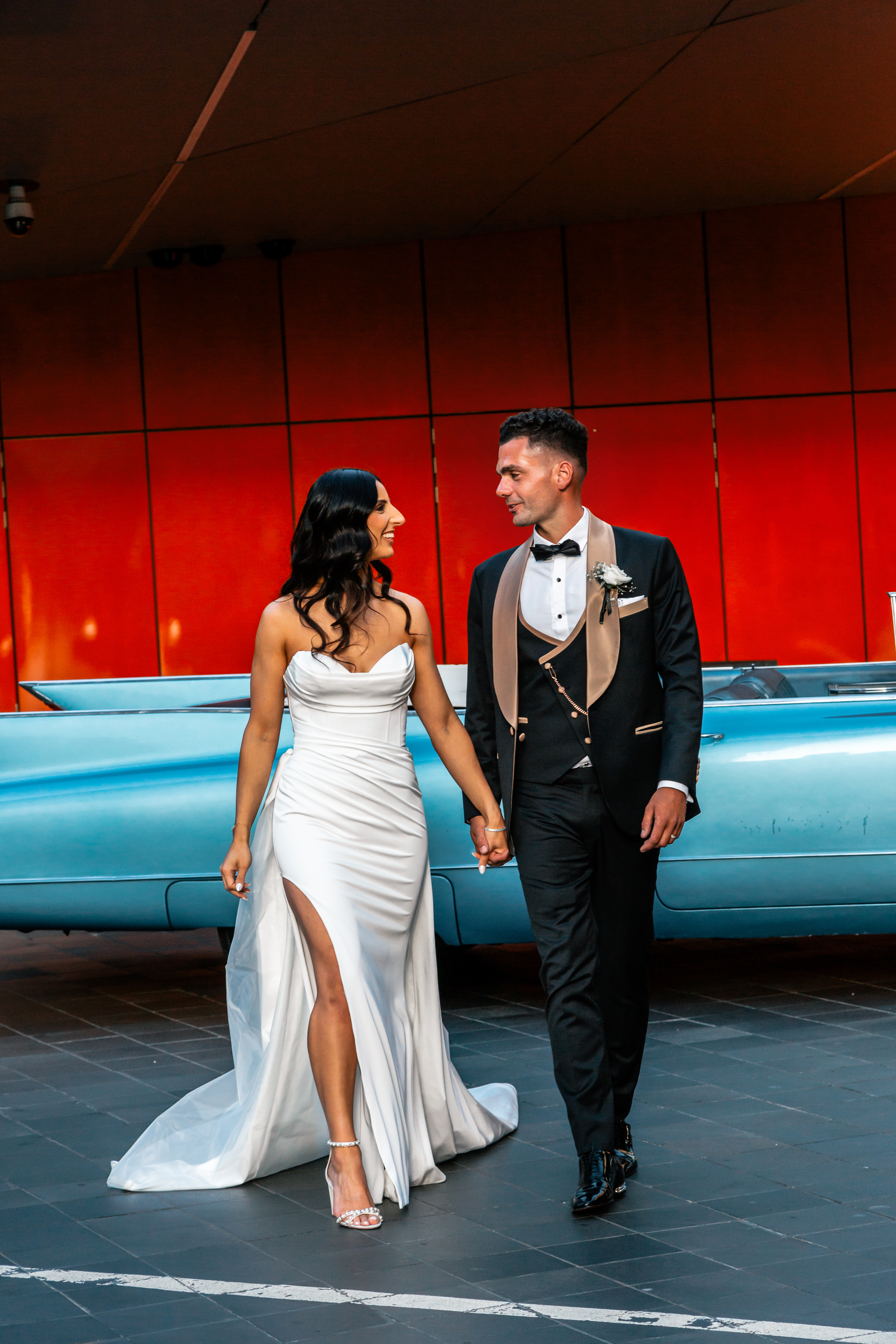 Romantic candid wedding photo of bride and groom walking hand-in-hand, smiling in front of a vintage blue car and bold red wall, natural fun wedding photography Melbourne, Yarra Valley, Mornington Peninsula