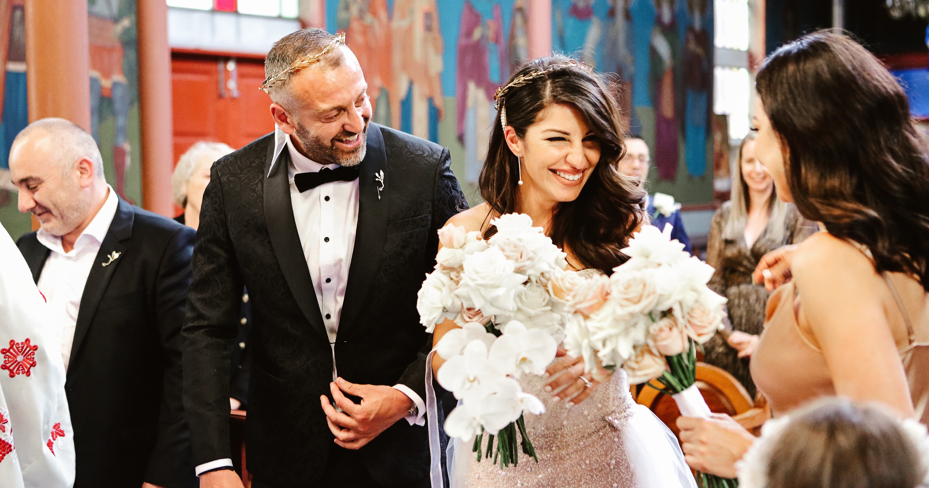 Candid photograph of a smiling bride and groom wearing traditional gold Stefana (crowns) during a traditional Greek Orthodox wedding ceremony in Melbourne. Authentic moment captured at the altar, featuring a modern white orchid and rose bridal bouquet