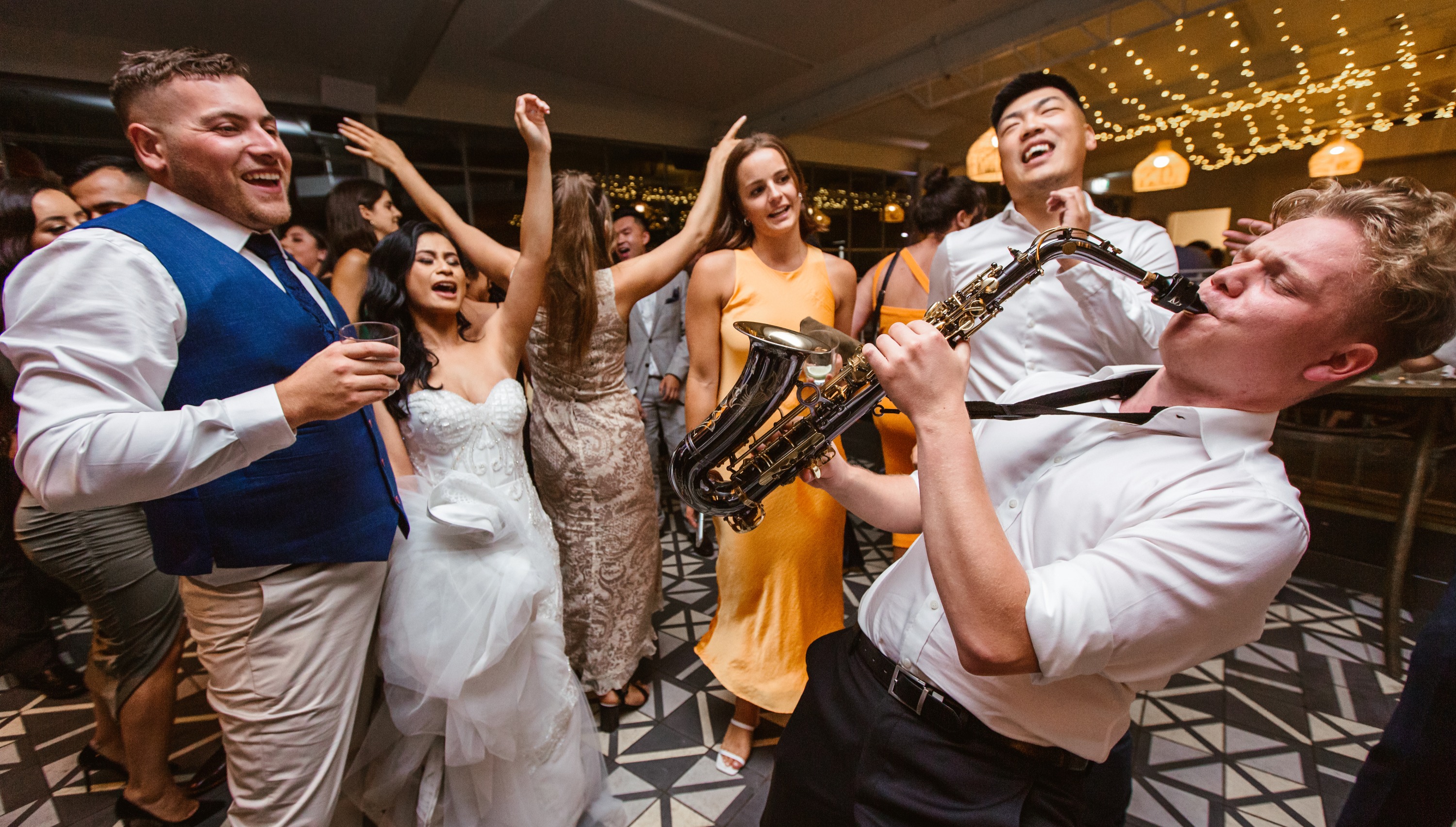 High-energy, candid photo of a saxophone player on a lively wedding reception dance floor surrounded by dancing and laughing guests. Modern inner Melbourne venue style with patterned floor and hanging fairylights, capturing a fun celebration
