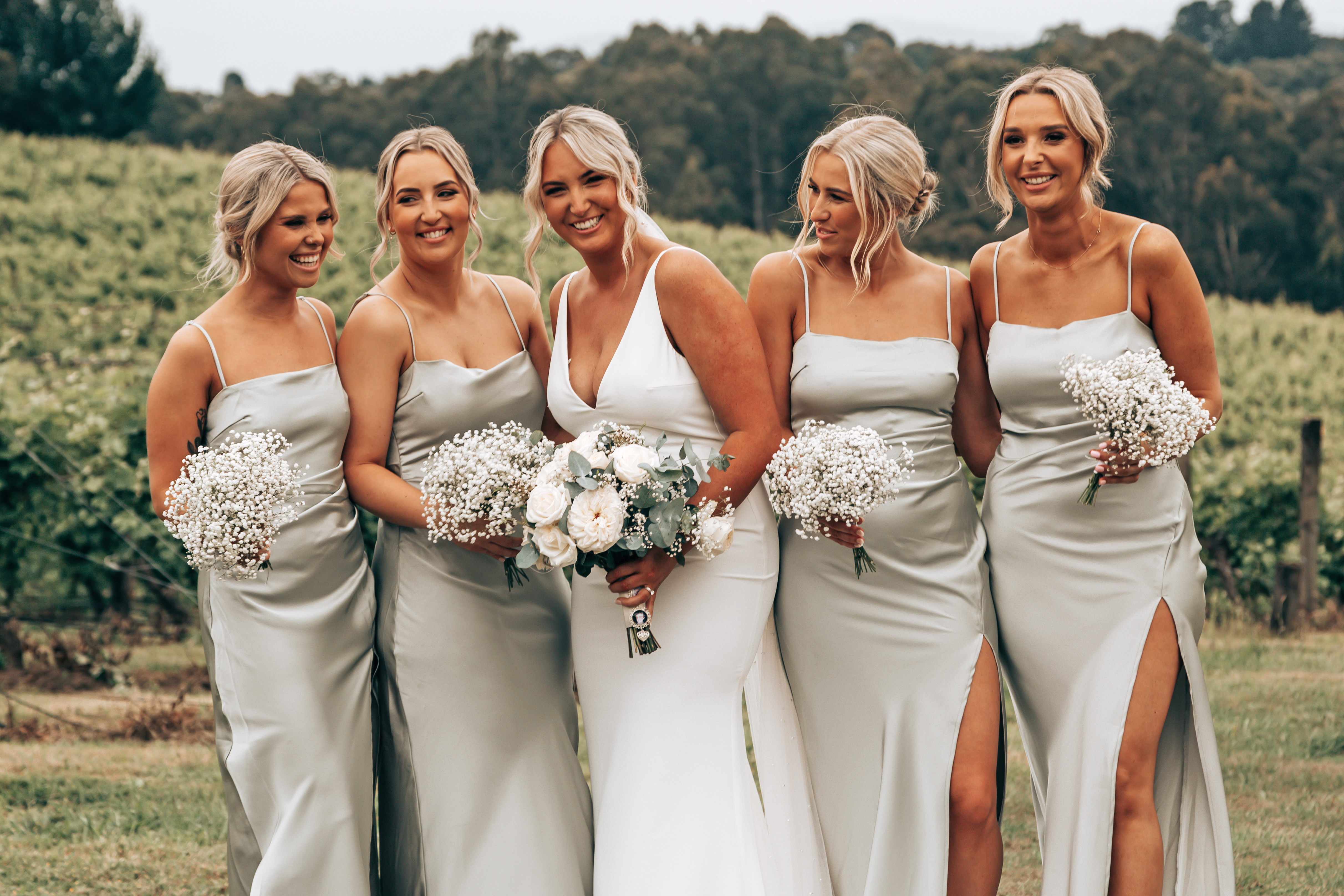 Bride in a white V-neck gown and four bridesmaids in matching sage green, high-leg-slit silk gowns, all holding baby's breath bouquets. Candid and unposed outdoor vineyard photo captured at a Yarra Valley winery