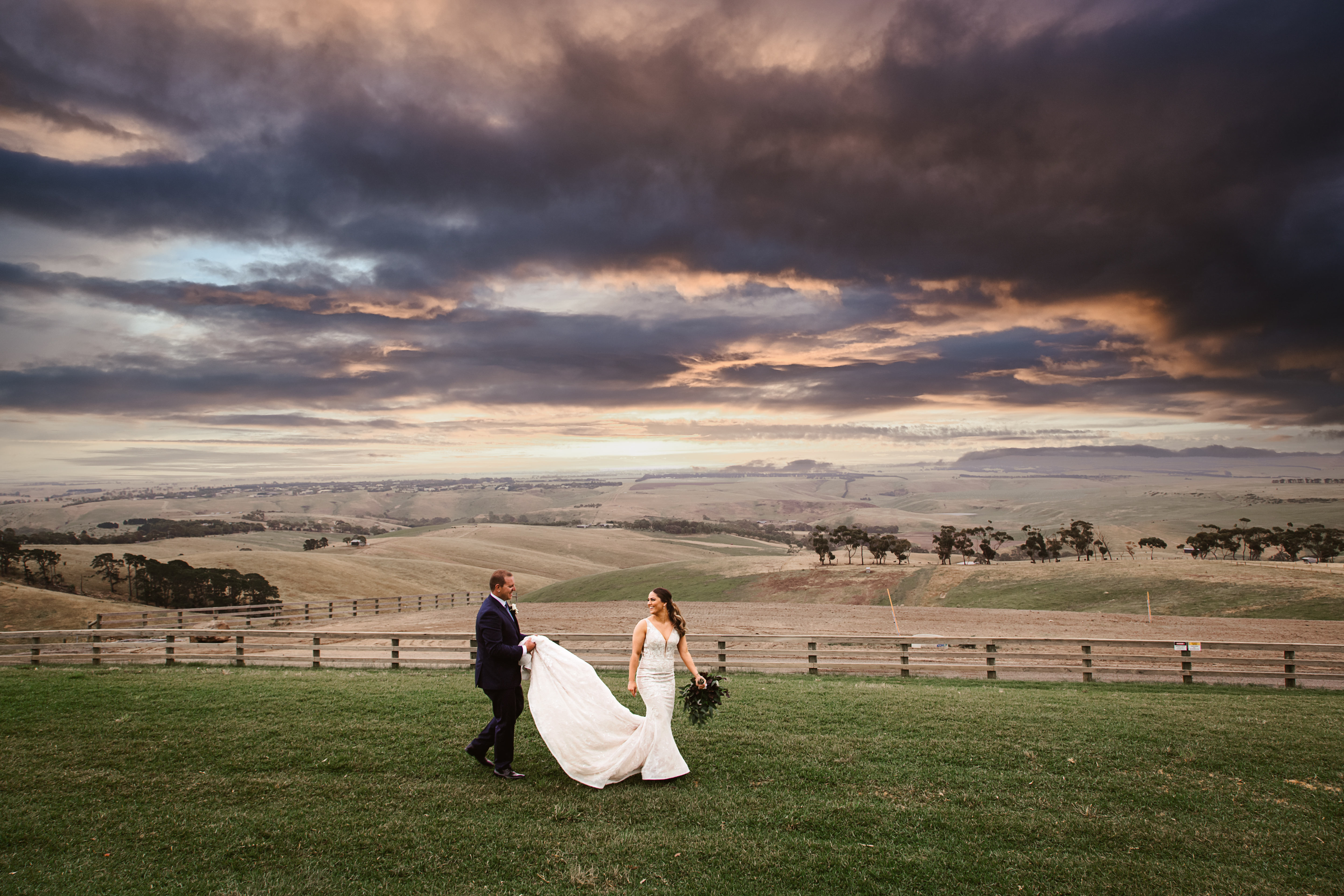 Dramatic wide-angle outdoor landscape wedding photograph. Bride and groom walking in a grassy field with rolling regional hills under a dark, dramatic sky. The groom is carrying the bride's train at the reception Marnong Estate Winery in Mickleham