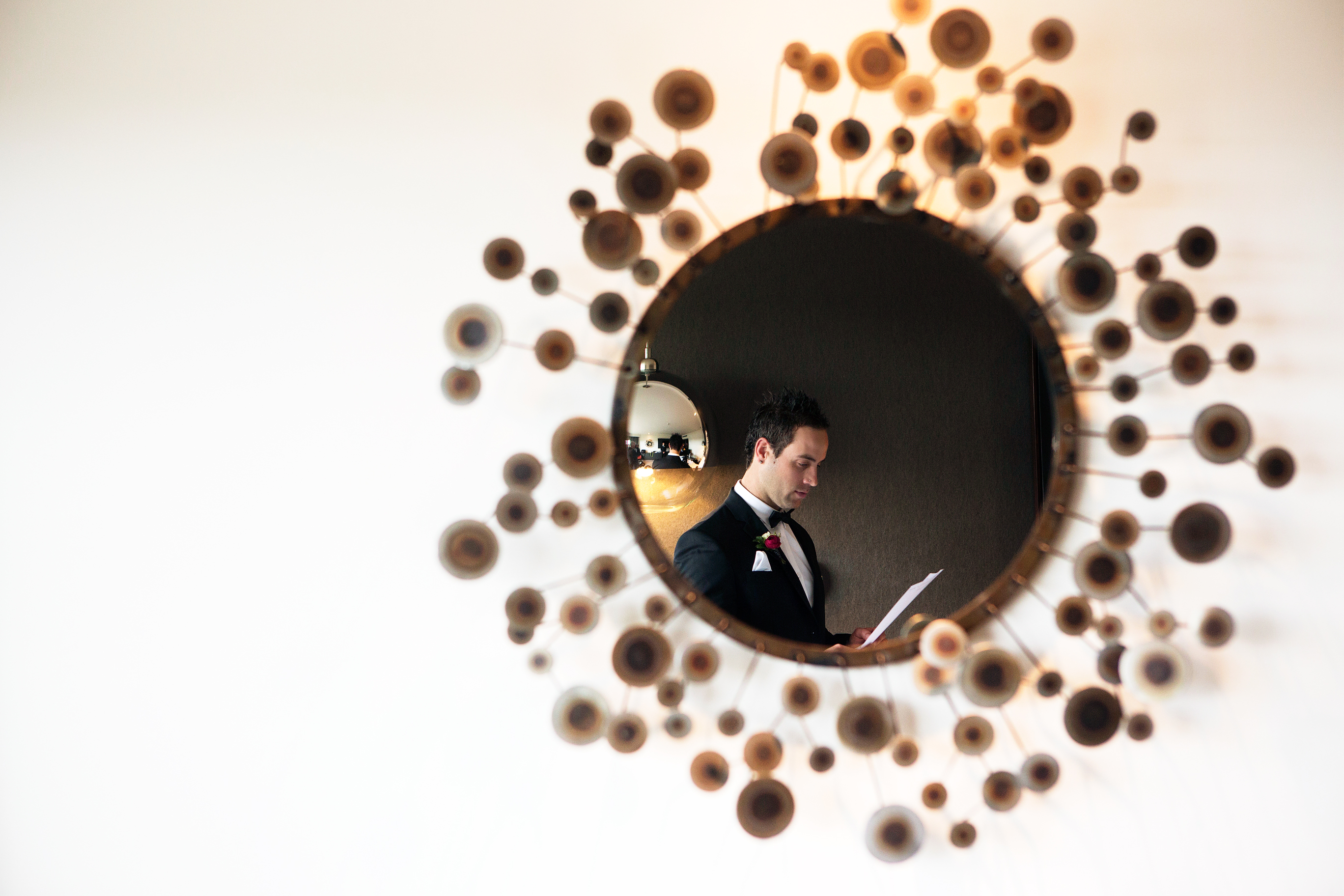 Artistic indoor wedding photo of a groom in a black tuxedo reading his vows. He is reflected in a geometric wall mirror composed of many circular brass discs, capturing an intimate and unposed moment during a Melbourne celebration