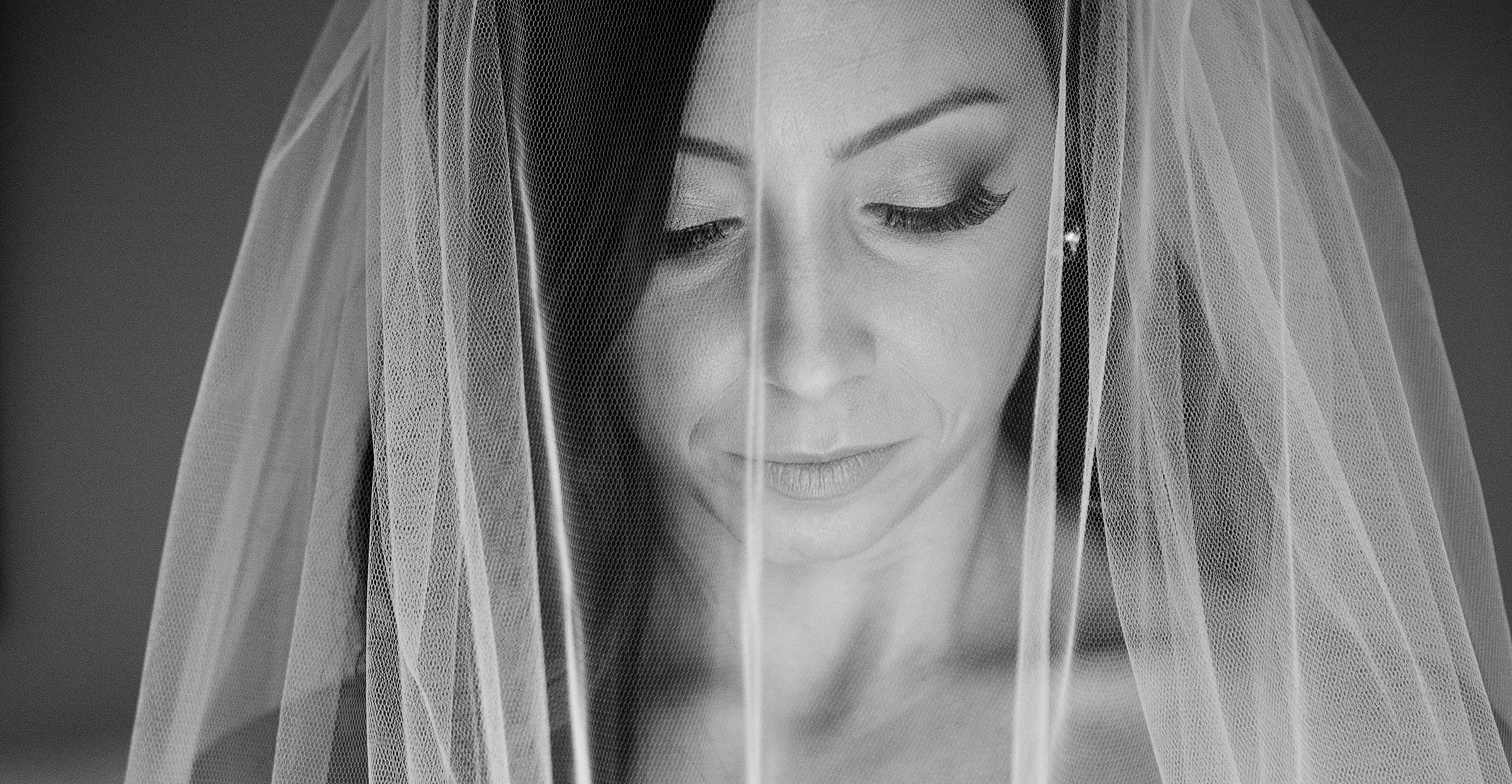 Tight, intimate close-up portrait of a bride's face viewed through a delicate lace veil. Artistic black and white photography focusing on specific details like smoky eye makeup and veil texture, perfect for modern editorial wedding inspiration